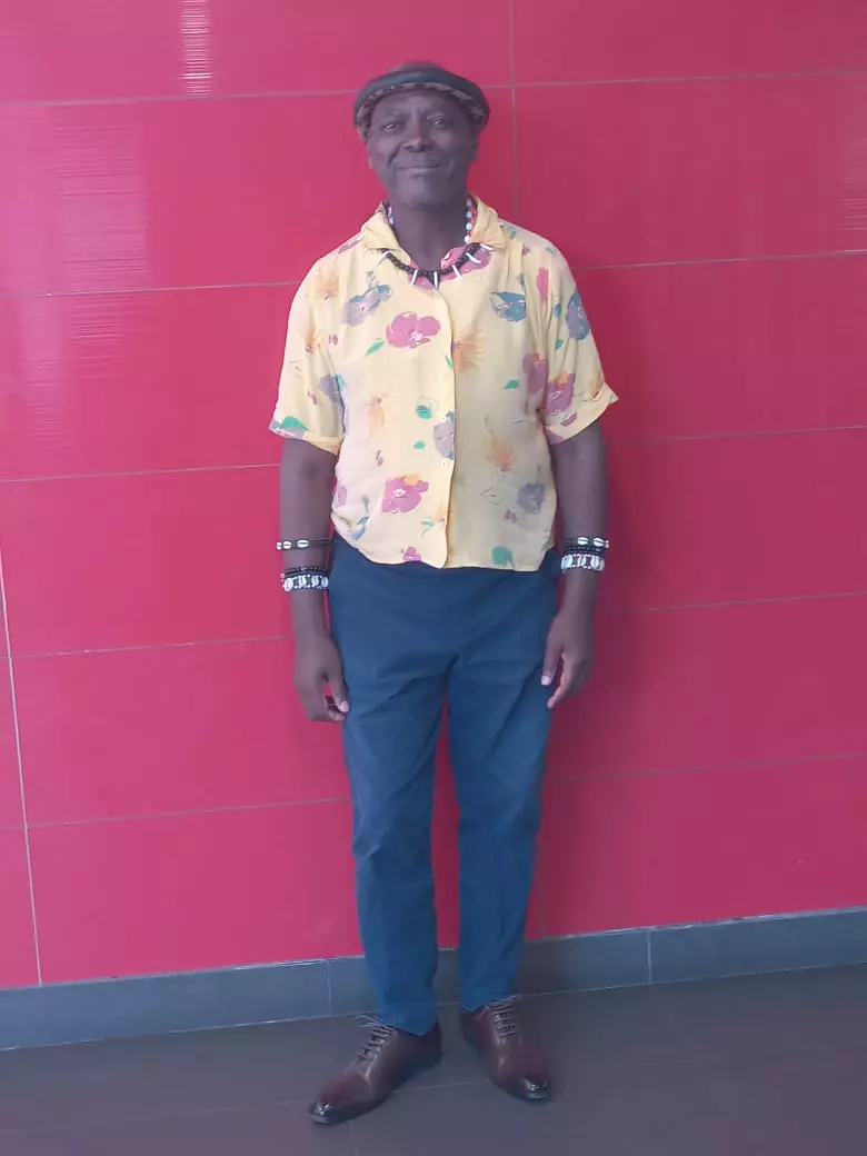 Emperor Thembu standing against a red tiled wall wearing a patterned yellow shirt and blue trousers