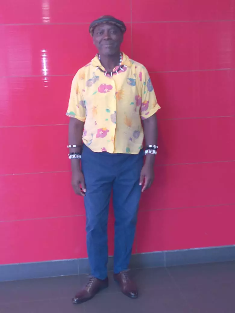 Emperor Thembu standing against a bright red tiled wall wearing a patterned yellow shirt and blue trousers